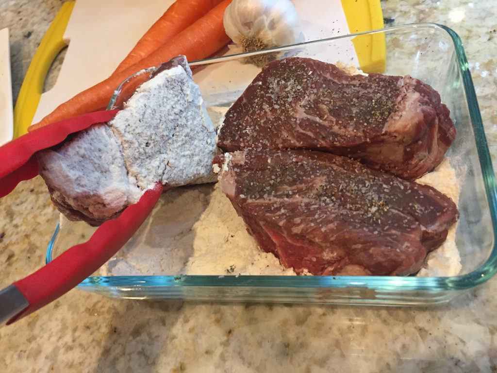 3 beef short ribs being dredged in flour in a rectangular glass dish. One coated in flour, the other two are not. 2 carrots and a head of garlic sitting on a white cutting board with