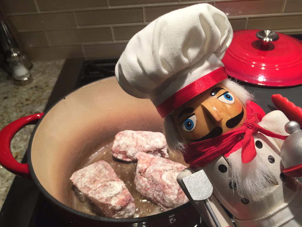 3 floured beef short ribs browning in a dutch oven with a nutcracker that looks like a chef.