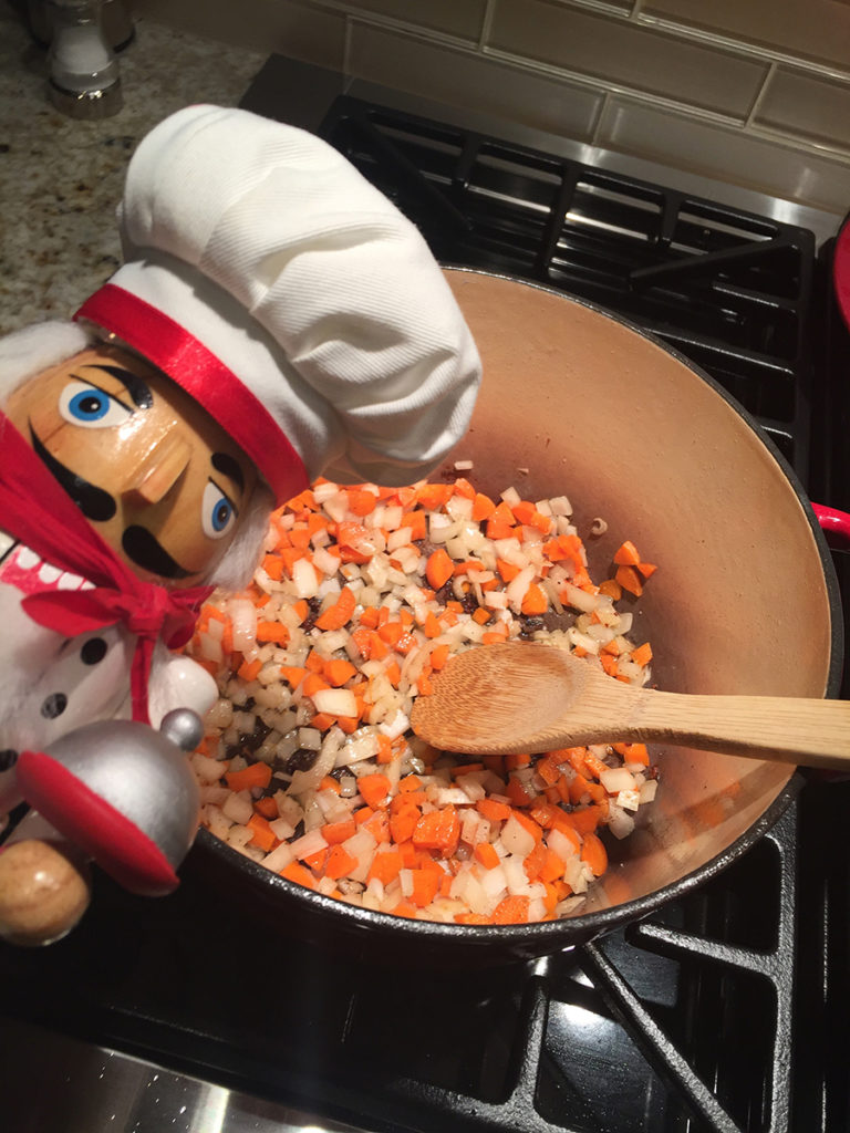 chopped carrots and onions in a dutch oven and stirred with a wooden spoon. There's a nutcracker that looks like a chef in the foreground.