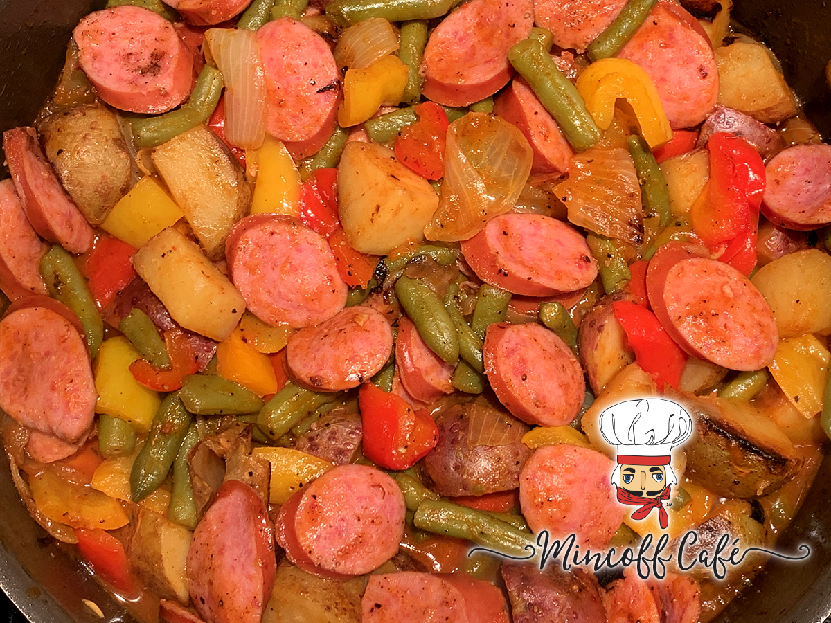 Skillet filled with sliced smoked sausage, onions, red and yellow bell peppers and green beans. There's a nutcracker in the foreground who looks like a chef. 
