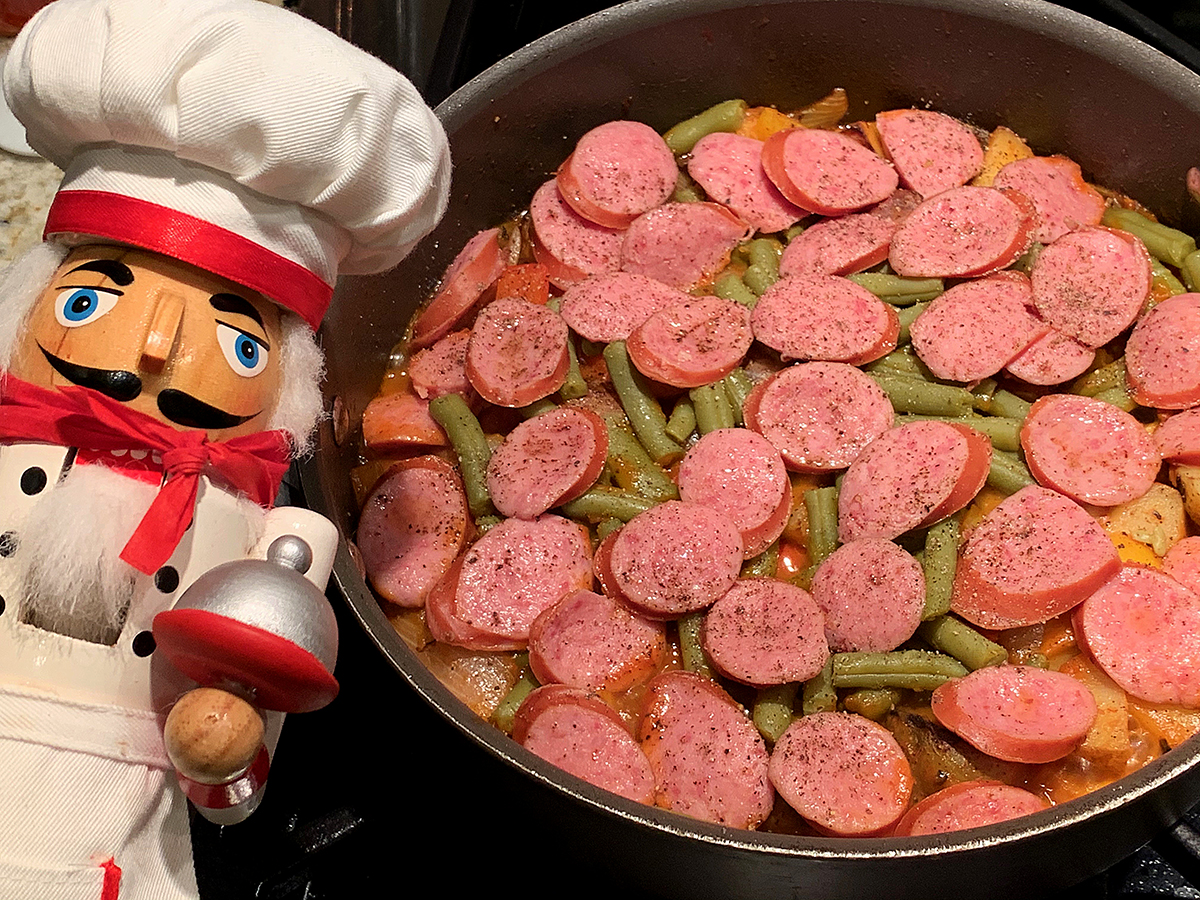 Skillet with green beans, pepper and potatoes with a layers of sliced polish sausage on top. There's a nutcracker in the foreground who looks like a chef. 