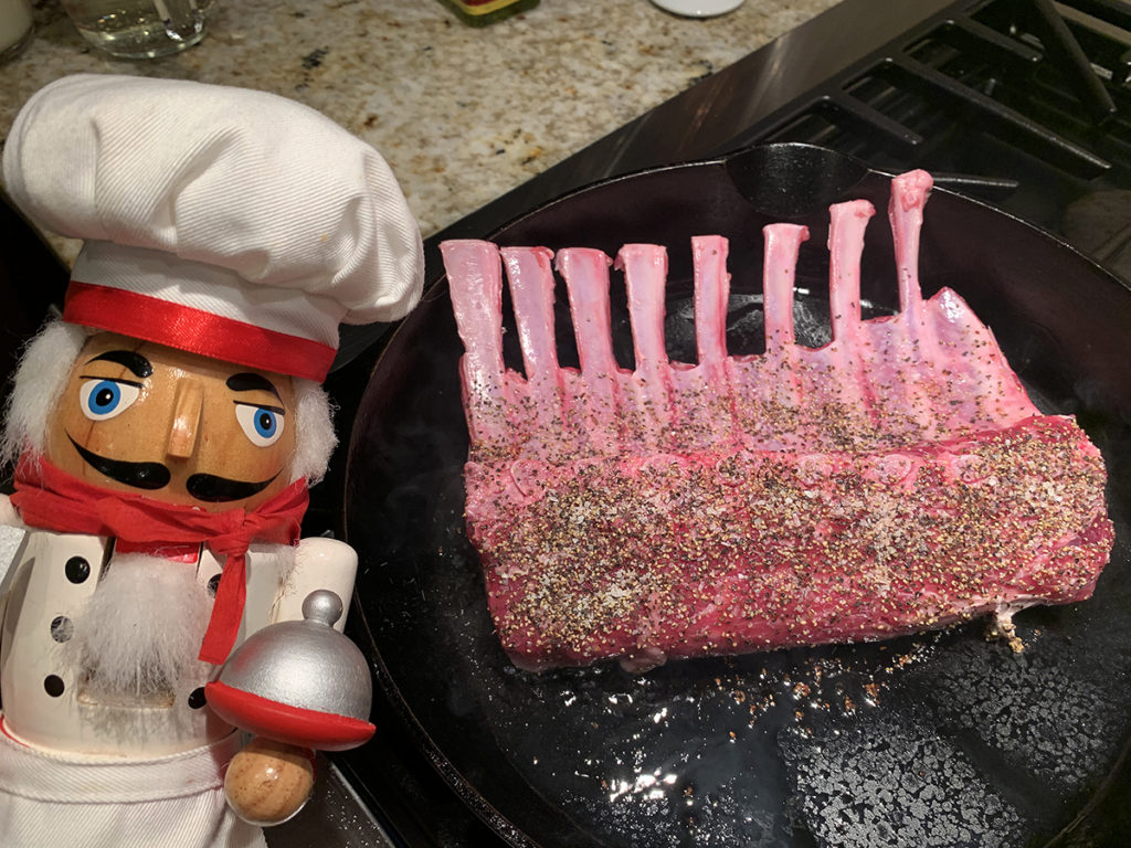 Raw rack of lamb being seared in an iron skillet. There's a nutcracker who looks like a chef in the foreground. 
