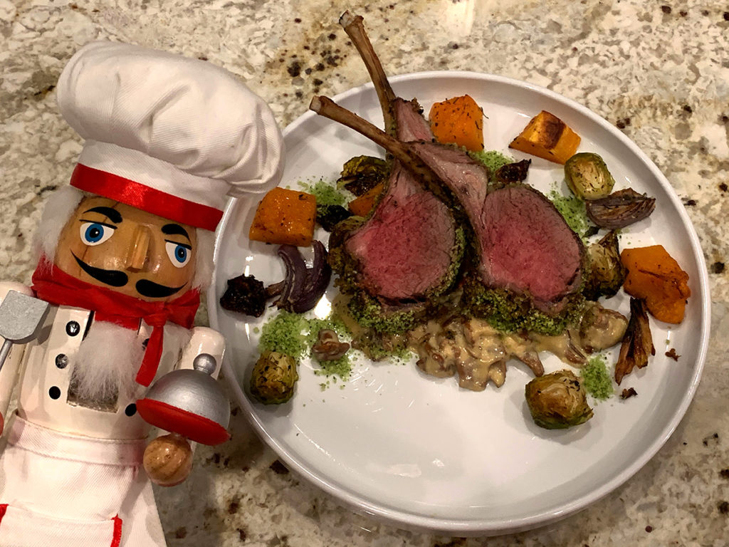 Two medium rare herb crusted racks of lamb with a chanterelle cream sauce on a round white plate with roasted butternut squash, brussels sprouts and red onions. All on a white round plate and with nutcracker who looks like a chef in the foreground.