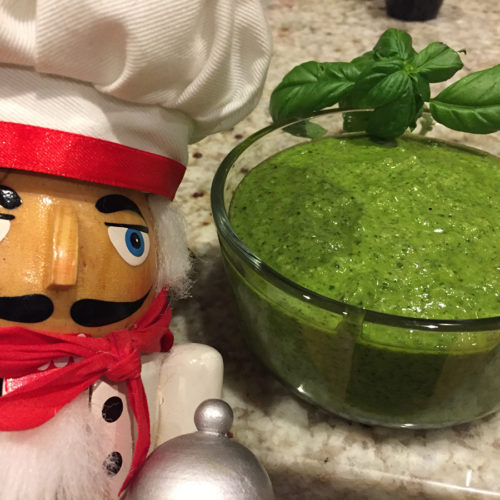 bright green homemade basil pesto in a clear glass bowl with a sprig of fresh basil on the rim of the bowl. There's a nutcracker in the foreground who looks like a chef.
