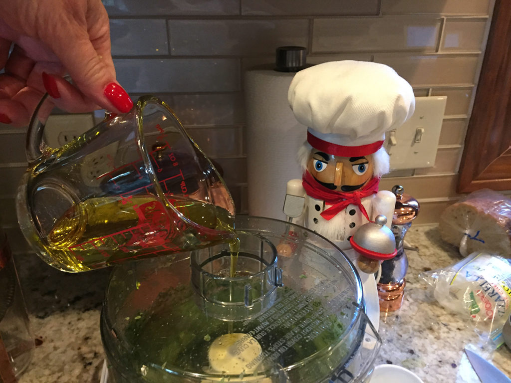 Adding the oil to the food processor to make fresh basil pesto. There's a nutcracker in the foreground who looks like a chef.