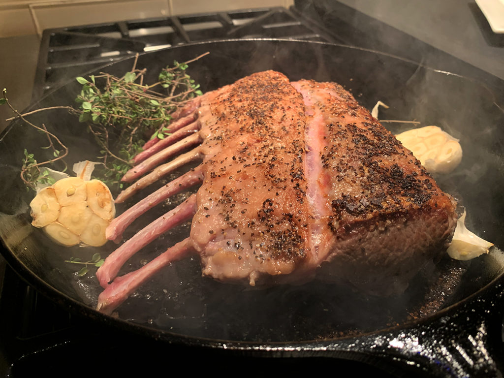 Seared rack of lamb with halved head of garlic and small bunch of thyme in an iron skillet. 