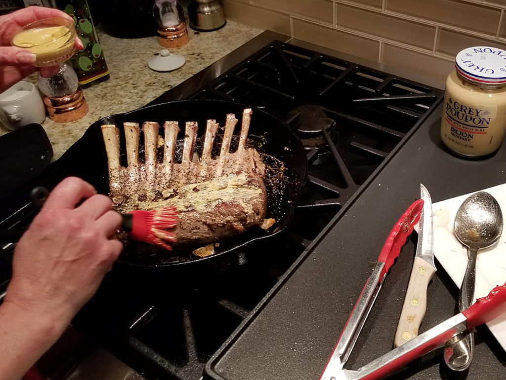 Seared rack of lamb in iron skillet getting brushed with dijon mustard. 