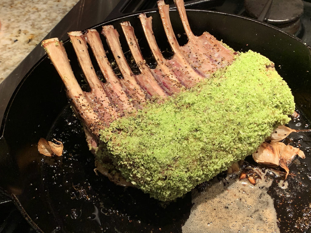 Roasted rack of lamb coated with a bright green herb breading in an iron skillet.