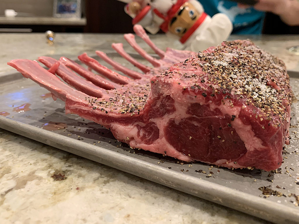 Raw rack of 8 lamb ribs on a sheet pan, heavily seasoned with salt and pepper. There's nutcracker who looks like a chef in the background. 