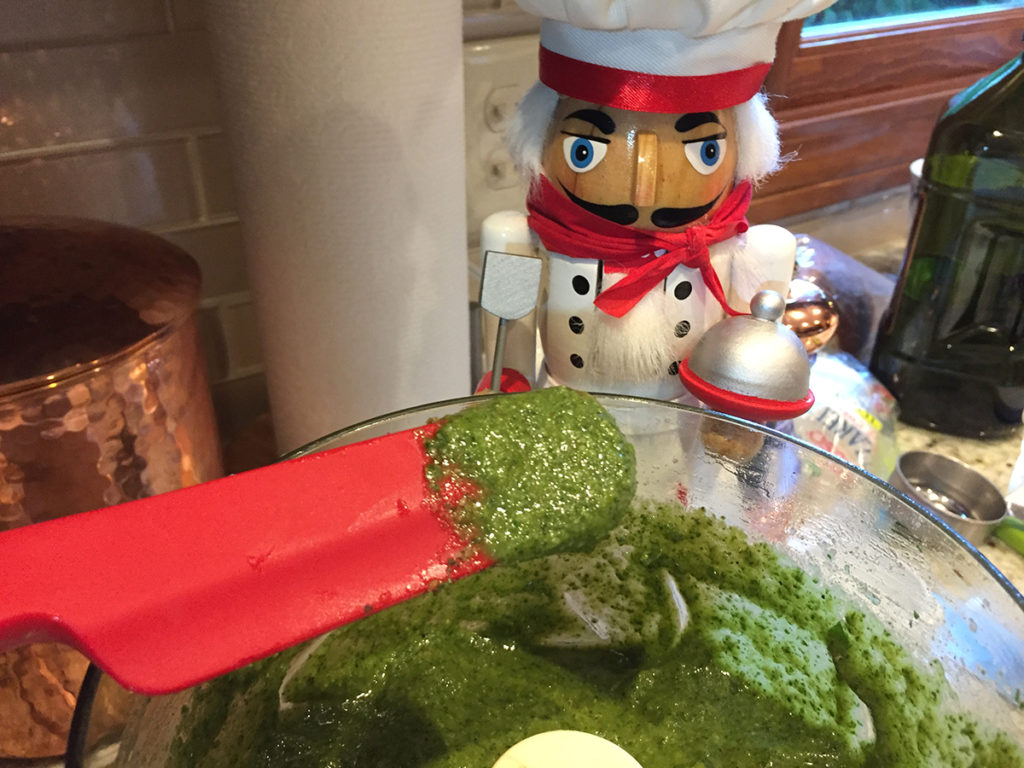 Pureed basil pesto in a food processor with a small amount on a red rubber spatula for tasting. There's a nutcracker in the foreground who looks like a chef. 