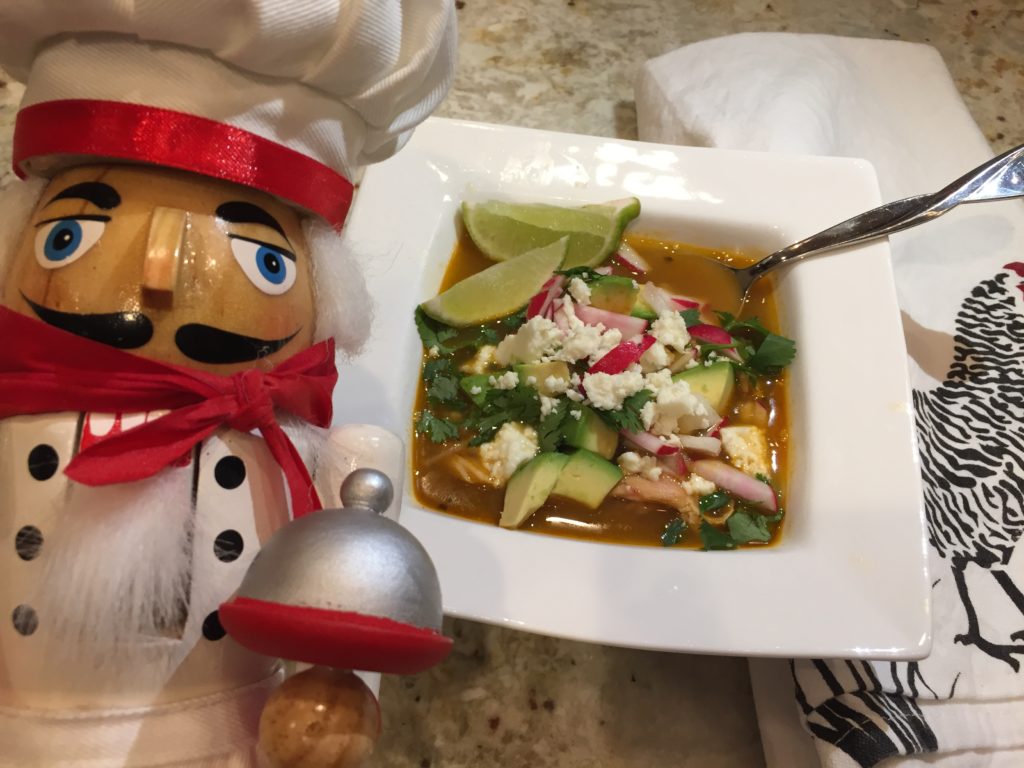 Chicken Posolé soup garnished with limes, red radishes, avocados, jalapeños and cilantro in a white square bowl. There's a nutcracker who looks like a chef in the foreground to the left.