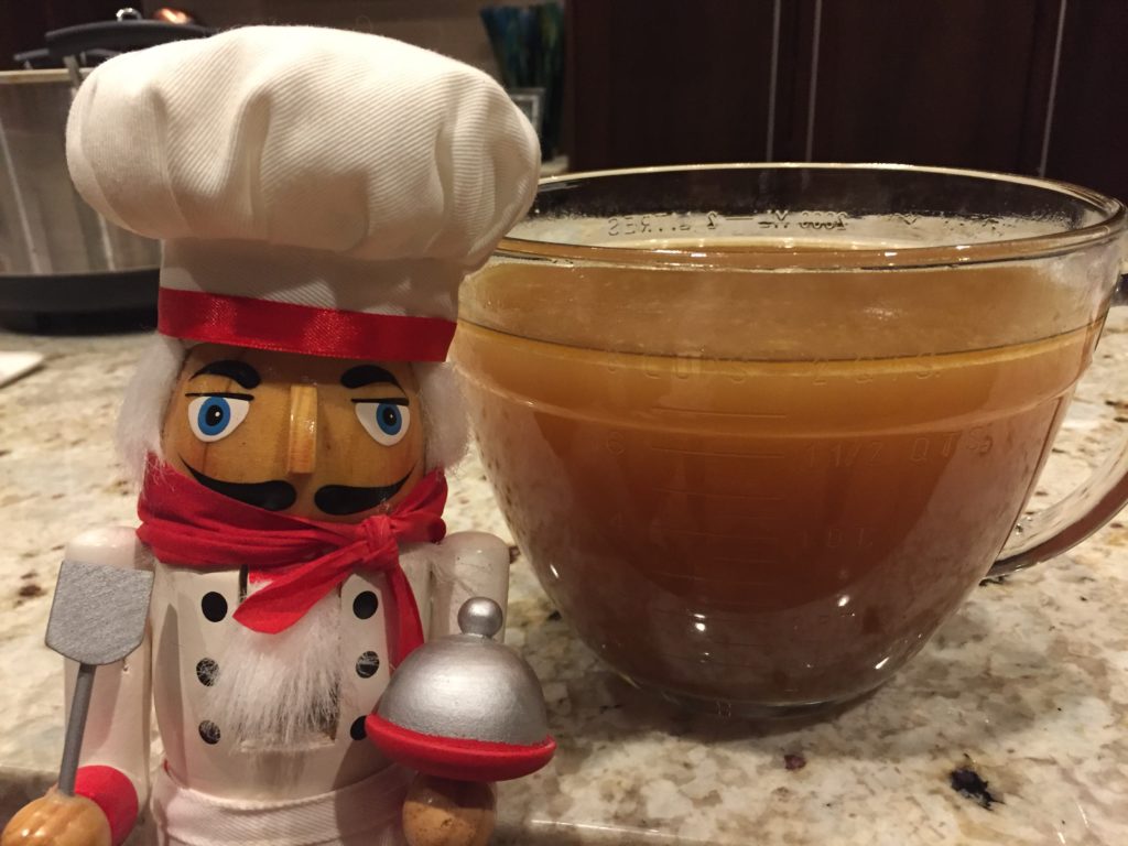 Large 8 cup glass measuring cup with brown chicken stock in it. It's setting on a granite counter top and there's a nutcracker who looks like a chef in the foreground on the left.