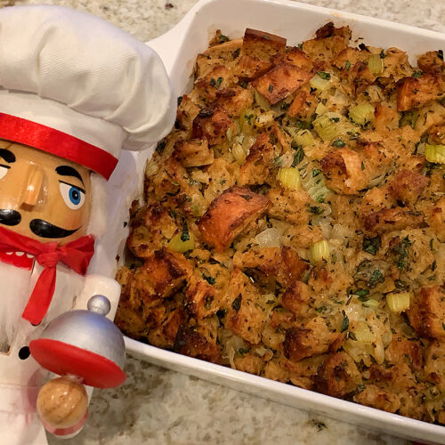 Square white dish filled with light golden brown cubes of bread and fresh herbs. There's a nutcracker in the foreground who looks like a nutcracker.