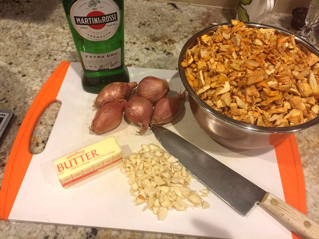 chopped chanterelles, chopped garlic, partial stick of butter, bottle of dry vermouth and a chef kife on a white cutting board