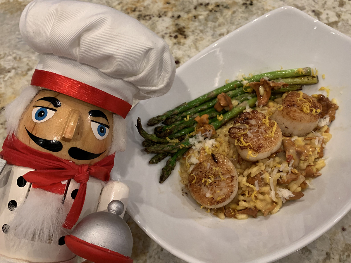 Seared scallops on a bed of a chanterelle and saffron risotto, sautéed asparagus in a white bowl with a nutcracker that looks like a chef