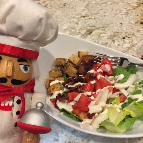 Pepe the nutcracker with a white bowl to the right of him containing a chopped salad consisting of bacon, lettuce, tomato, onion, croutons and ranch dressing