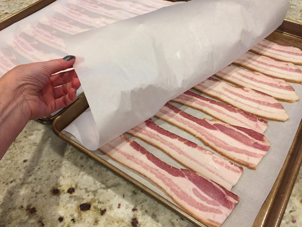 raw bacon sandwiched between layers of parchment paper on baking sheet. female hand pulling corner of parchment open to show strips of bacon