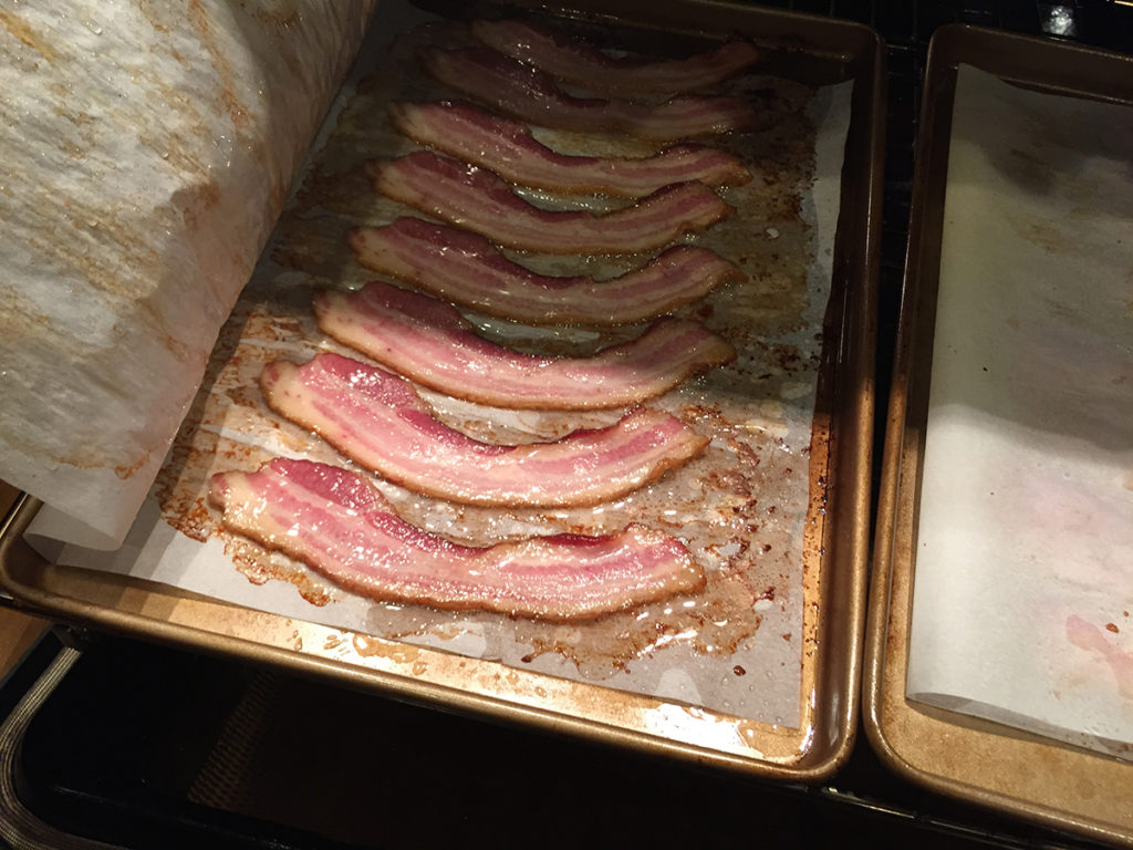 Partially cooked bacon on baking sheet, sandwiched between layers of parchment paper. Paper pulled away to show bacon