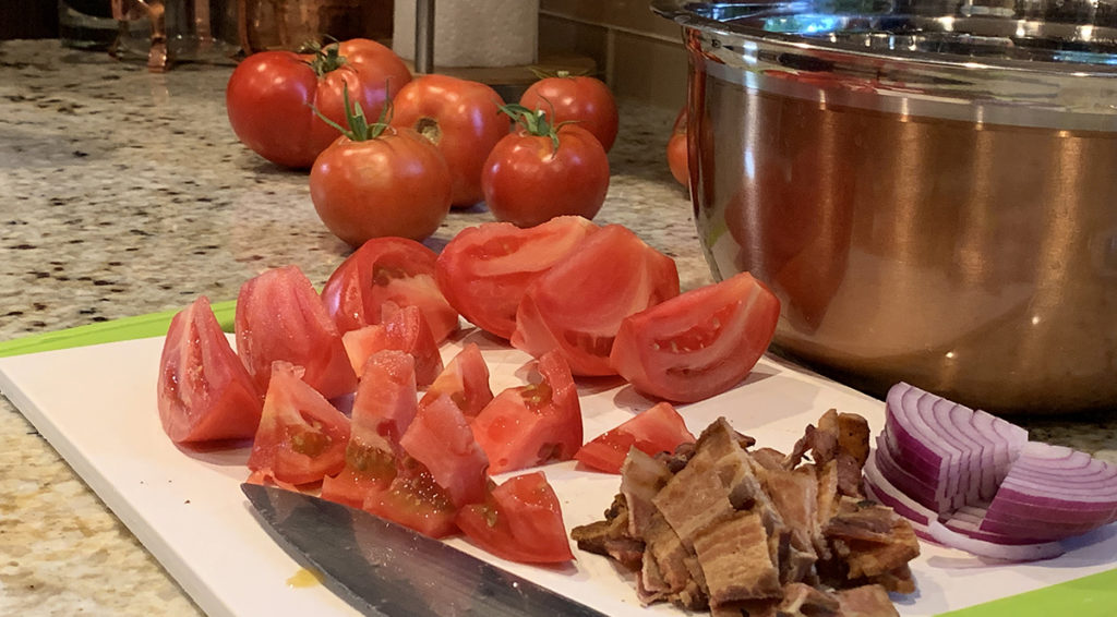 Several whole tomatoes in background with more cut up tomatoes in foreground on cutting board. Also has chopped cooked bacon on cutting board with knife. Brass bowl in background on the right. 