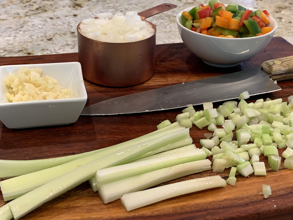 Chopped garlic, onions and tri colored bell peppers portioned into separate bowls, along with celery being chopped. all on a wood board with a chef knife. 