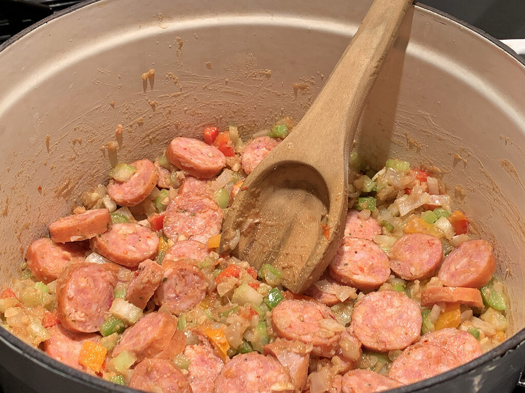 Slices of pinkish sausage added to the white dutch oven on top of the onions, peppers and celery. 