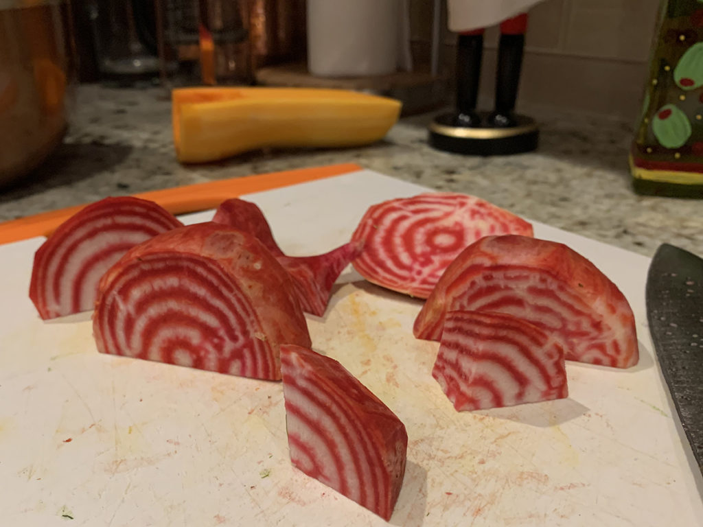 Candy cane beet root peeled and chopped showing it's red and white spirals. They are on a white cutting board with a chef knife and you can see a half of a butternut squash in the background.