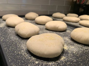 close up of partially cooked, and very puffy homemade English muffins on cornmeal sprinkled griddles.