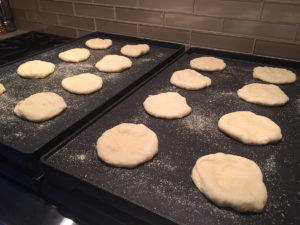16 Raw round pieces of dough on two cornmeal sprinkled griddles.
