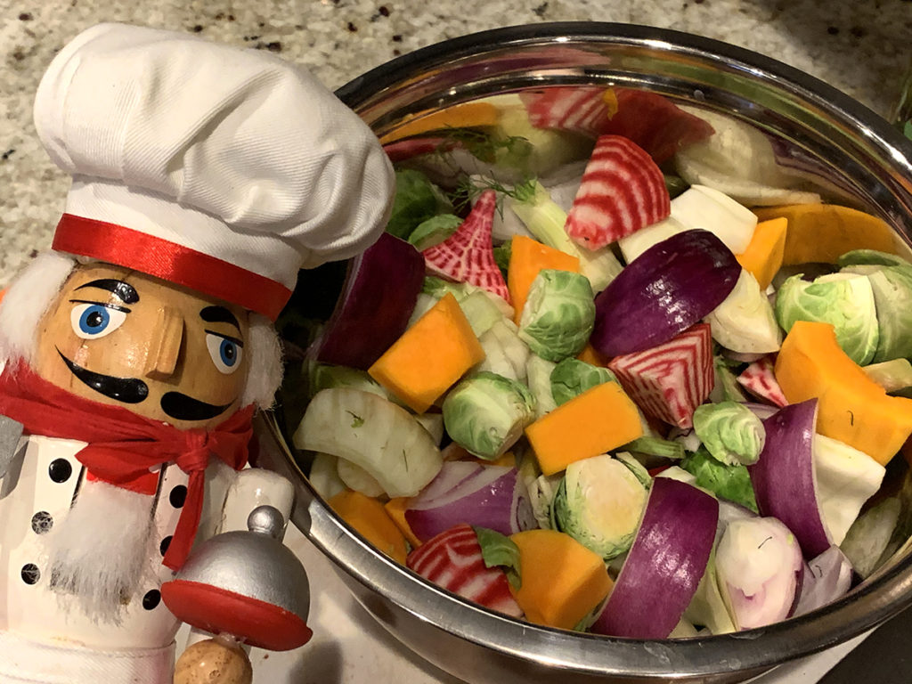 A stainless steel bowl of colorful, mixed, fall vegetables (butternut squash, red onions, brussles sprouts, fennel bulb and candy cane beet root) with a nutcracker who looks like a chef in the left foreground.