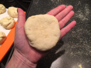 3.5" piece of flattened dough in the palm of a woman's hand. Griddle with sprinkled cornmeal below and balls of dough ready to be flattened to the left.