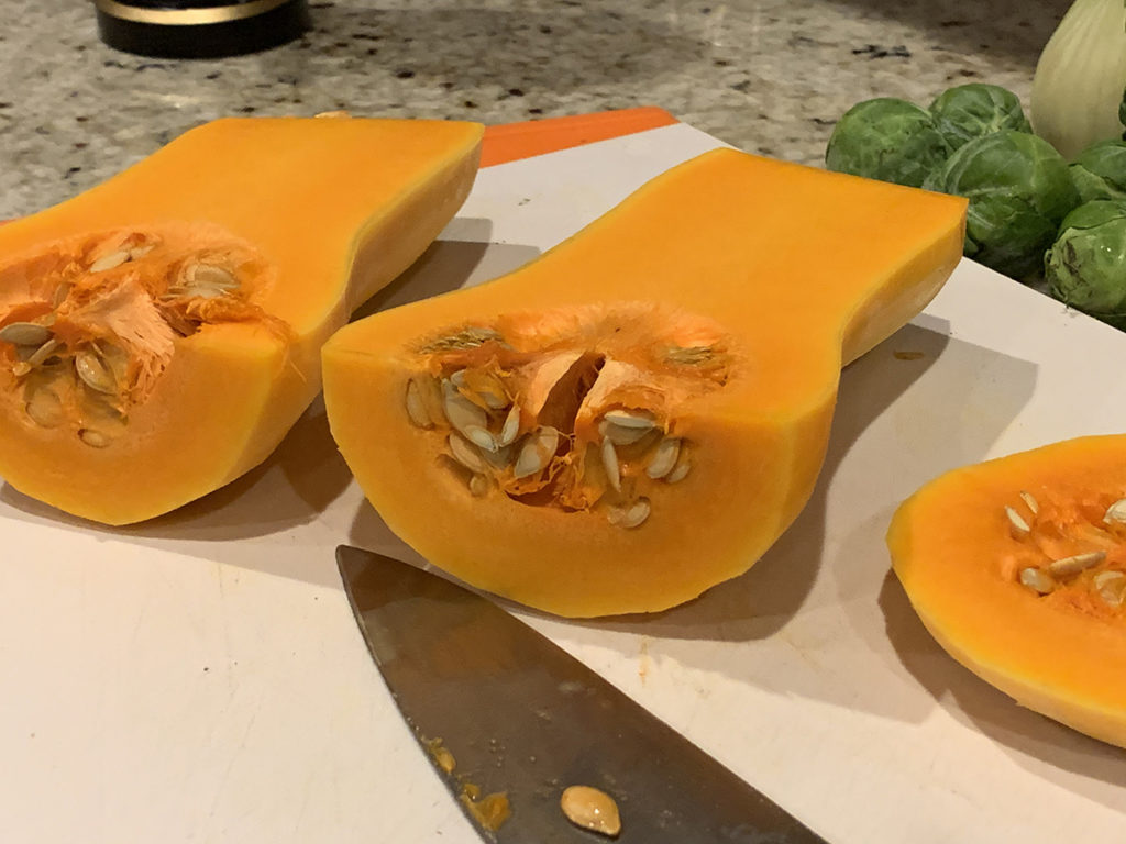 butternut squasg peeled and halved. Seeds are still in and both halves are sitting on a white cutting board with a chef knife in front. 