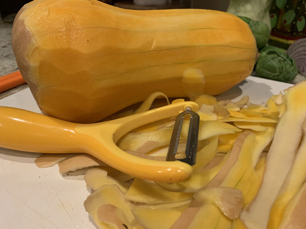 Peeled butternut squash with peelings and the peeler laying in front of it and all on a white cutting board. 