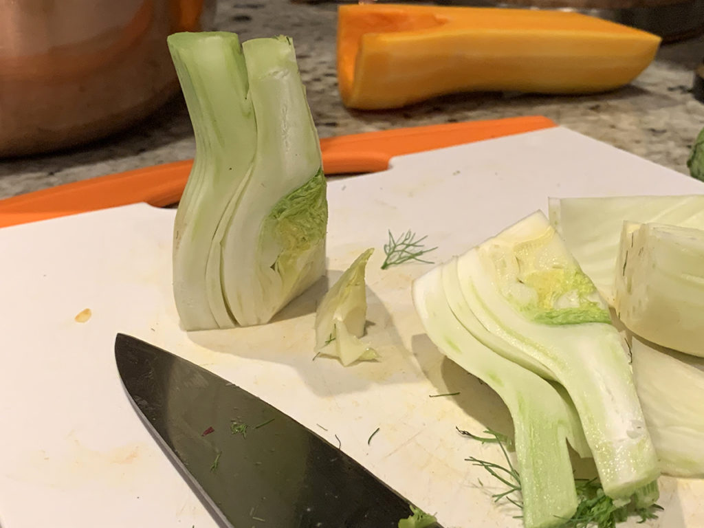 two quarters of a fennel bulb sitting on a white cutting board. One has the core removed. There's chef knife in the foreground and a butternut squash half in the background.