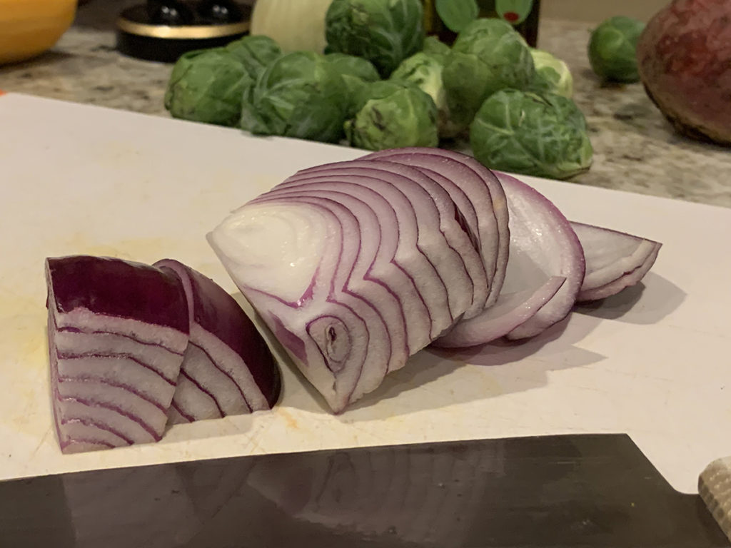 large chunks of red onions on a white cutting board with a chef knife in the foreground and some uncut brussles sprouts in the background.