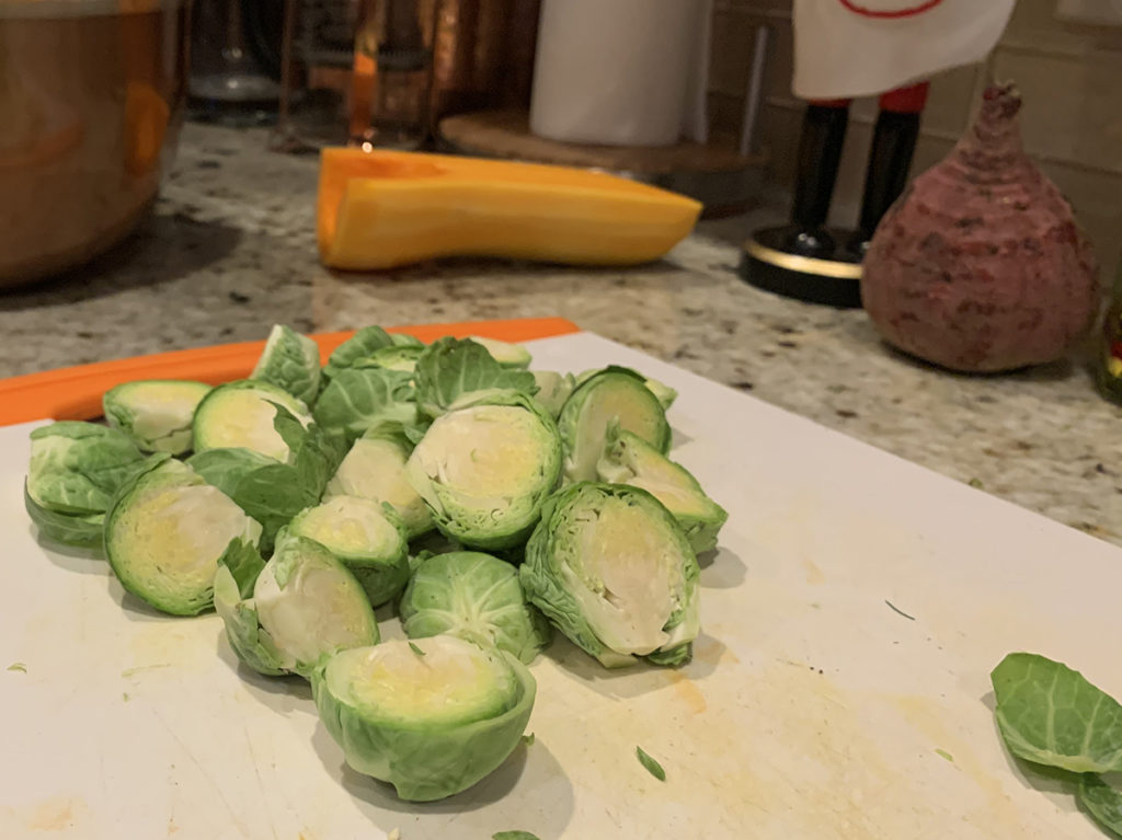 a small pile of Brussels sprouts cut into halves on a white cutting board. Half of a butternut squash and a beet root in the background.