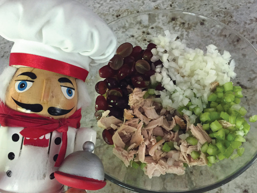 Chopped smoked turkey, celery, onions and red grapes in a clear glass bowl with a nutcracker that looks like a chef in the foreground to the left.