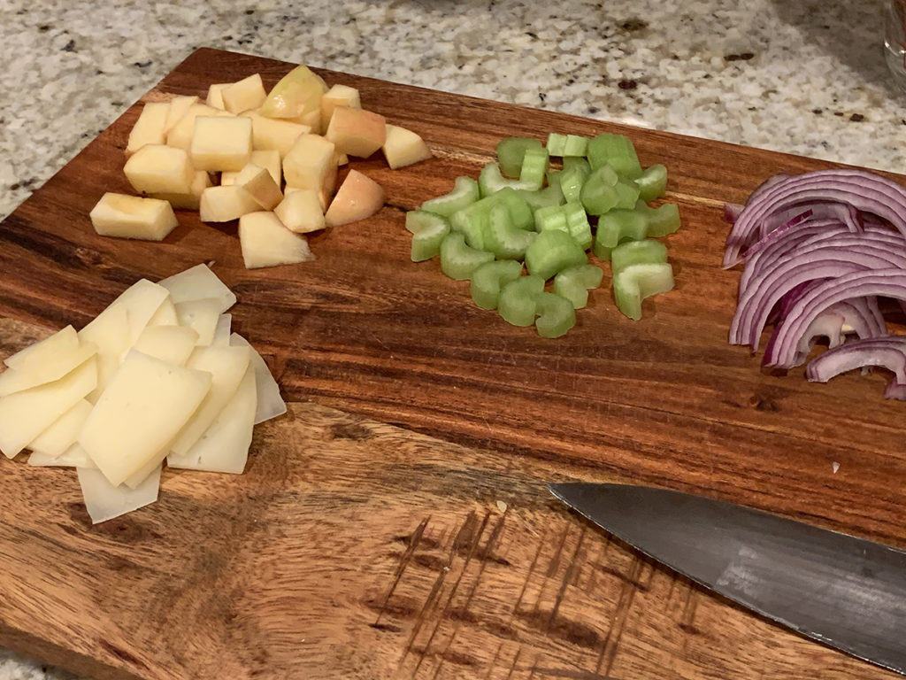 individual piles of chopped apple, chopped celery, small slices of Manchego cheese, and slices of red onions, all on a wood cutting board with a chef knife. 