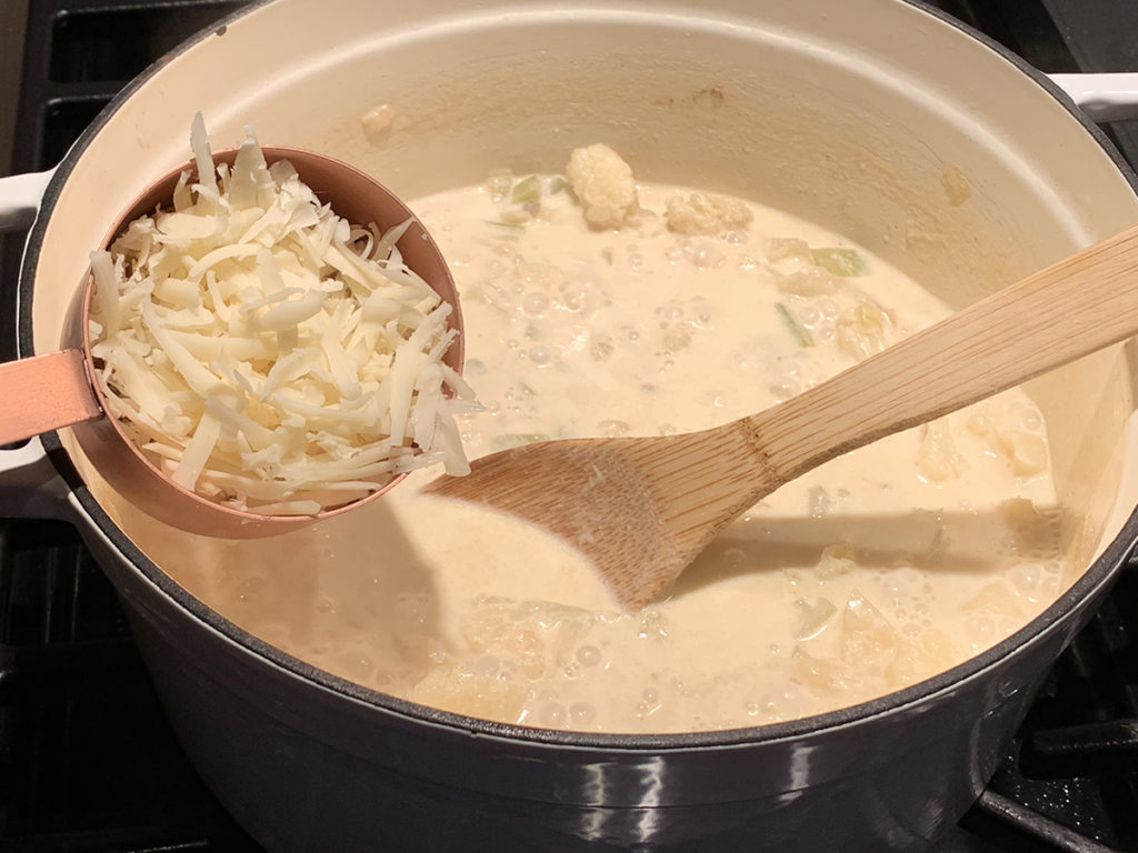 Pale yellow cauliflower soup in a white dutch oven with a wooden spoon, and a copper measuring cup filled with Gruyere cheese ready to pour in.