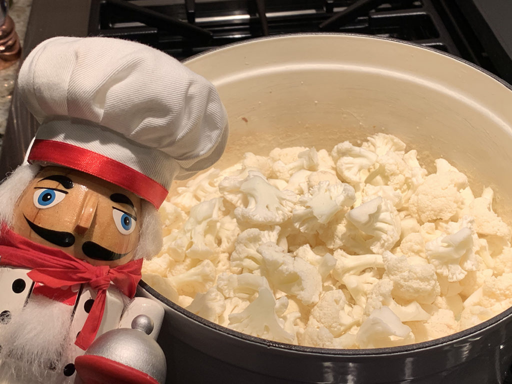 Chunks of cauliflower added to a white dutch oven with a nutcracker who looks life a chef in the foreground on the left.