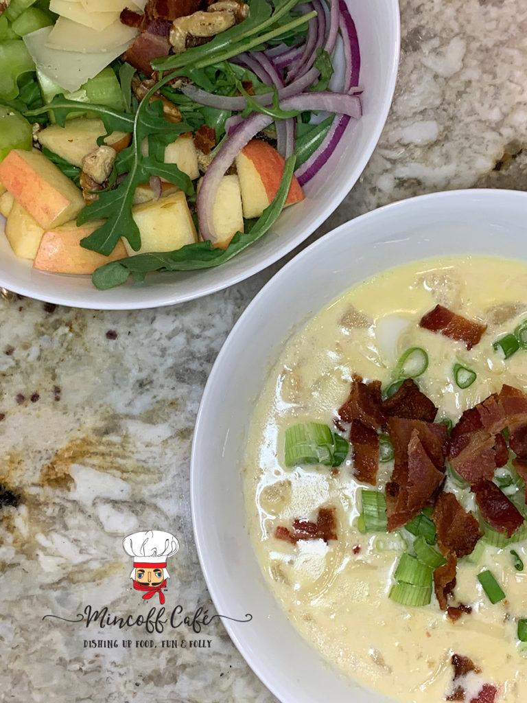 Vertical image from overhead of a salad containing bacon, apple, arugala, walnuts, and red onions in the upper left corner, and a bowl with cauliflower soup garnished with green onions and bacon in the bottom right corner. 