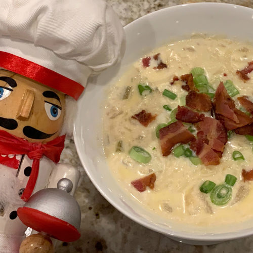 Pale yellow cauliflower soup in a white bowl, garnished with chopped green onions and bacon. A nutcracker that looks like a chef in the foreground.