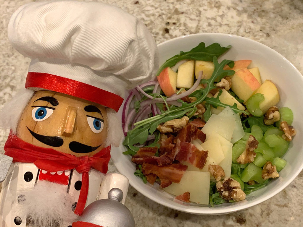 Bacon, Apple, celery, red onions, walnuts, manchego cheese and arugala salad in a while bowl, with a nutcracker who looks like a chef in the foreground on the left.