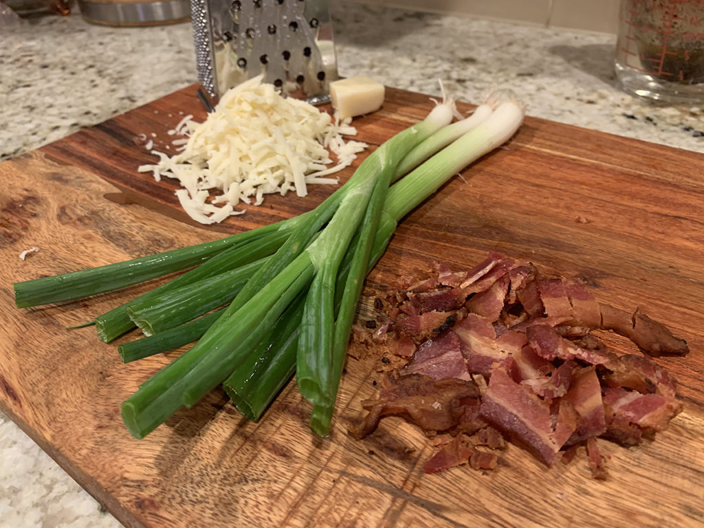 chopped bacon, whole green onions and shredded gruyere cheese on a wood cutting board