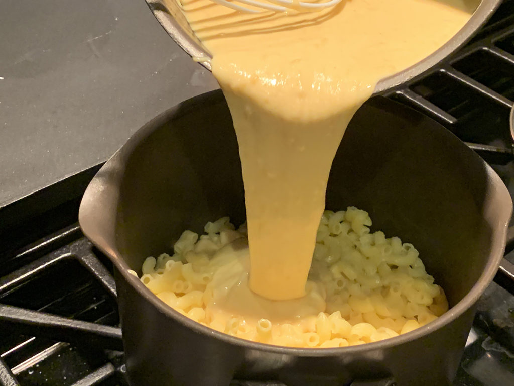 Pale yellow cheese sauce being poured into pan containing gluten free elbow pasta. 