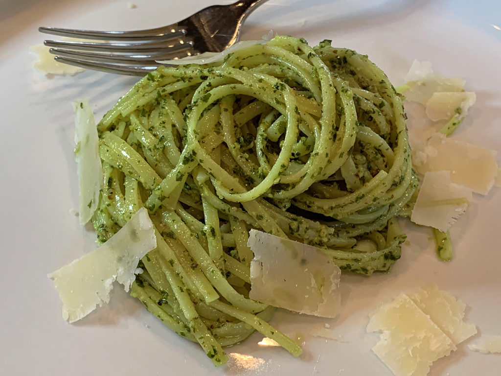 A swirled pile of linguini coated with green stinging nettle pesto sauce and a few shavings of parmesan cheese on a white plate with a fork.