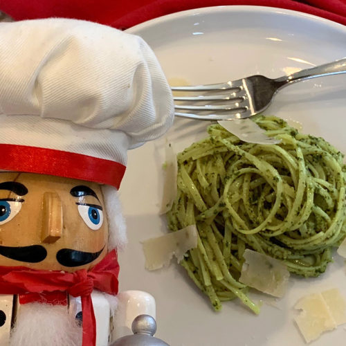 A swirled pile of linguini coated with green stinging nettle pesto sauce and a few shavings of parmesan cheese on a white plate with a fork. And a nutcracker who looks like a chef in the foreground.