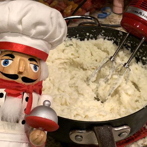 Mashed potatoes being made in a large sauce pan with a hand mixer. There's a nutcracker that looks like a chef in the foreground.