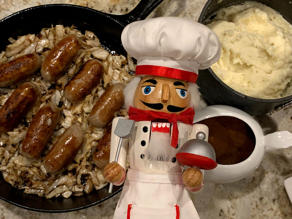 Overhead view of Irish style sausages (bangers) in an iron skillet, mashed potatoes (mash) in a saucepan, a small white gravy boat filled with onion gravy and a nutcracker who looks like a chef in the center.