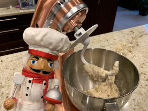 Copper colored KitchenAid mixer kneading pizza dough with dough hook. There's a nutcracker who looks like a chef in the foreground.