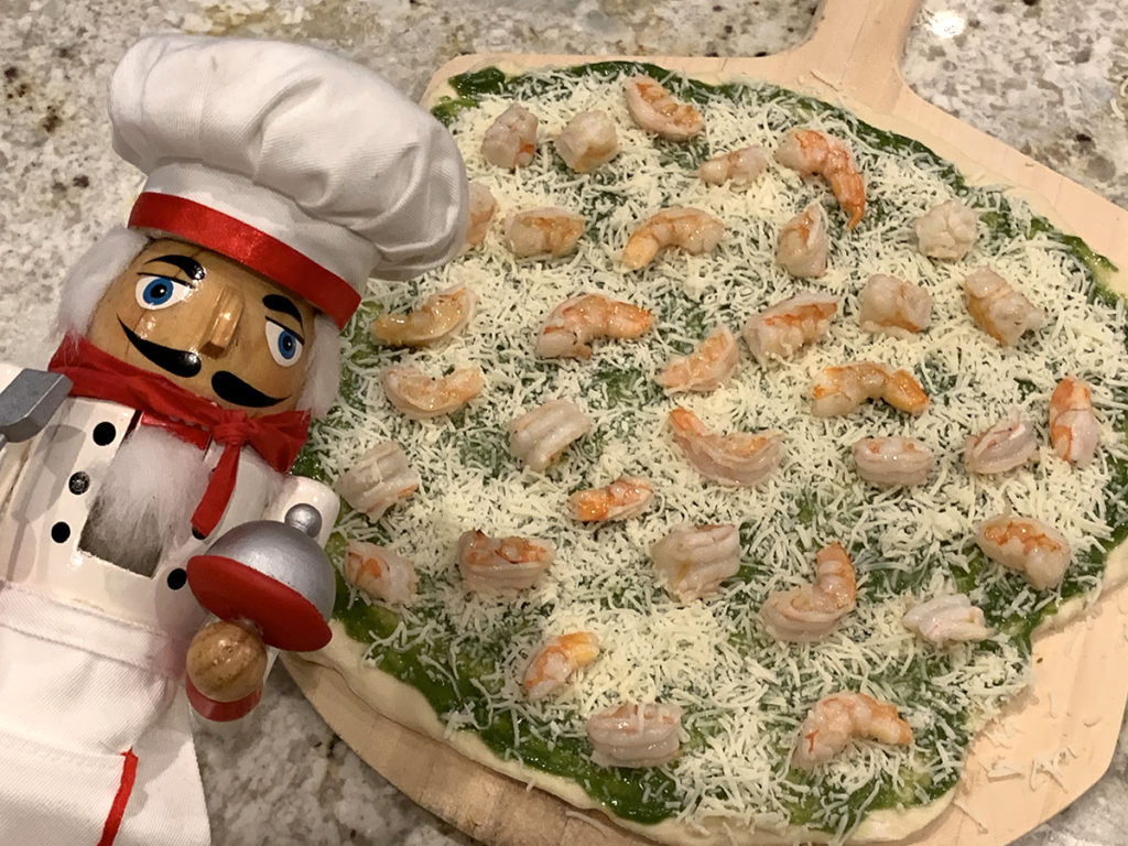 Raw shrimp & nettle pesto pizza topped with fresh arugula and parmesan cheese on a wood pizza peel. There's a nutcracker who looks like a chef in the foreground.